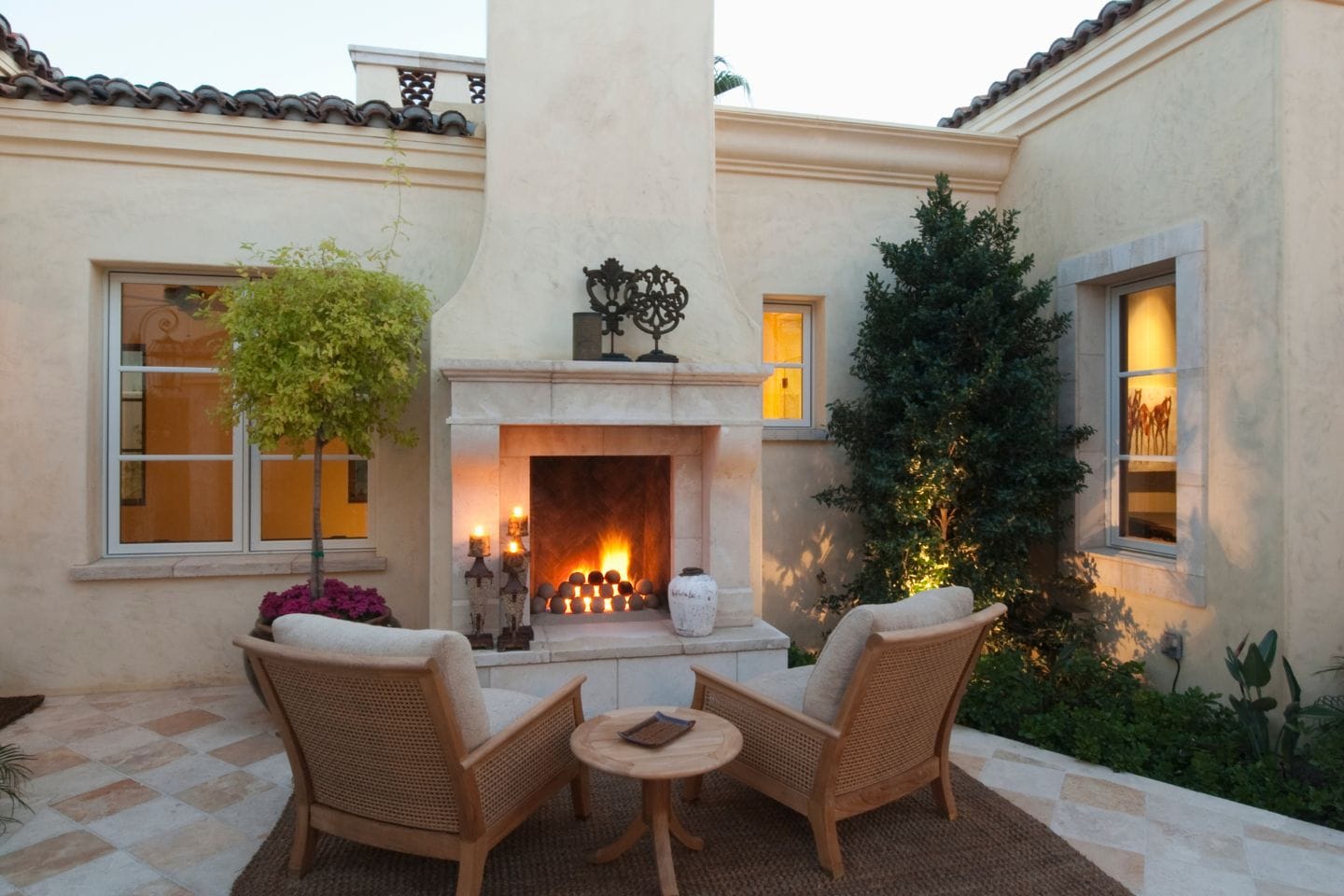 Outdoor patio with a glowing outdoor fireplace, two wicker chairs, a small round table, and surrounding potted plants against a stucco building.