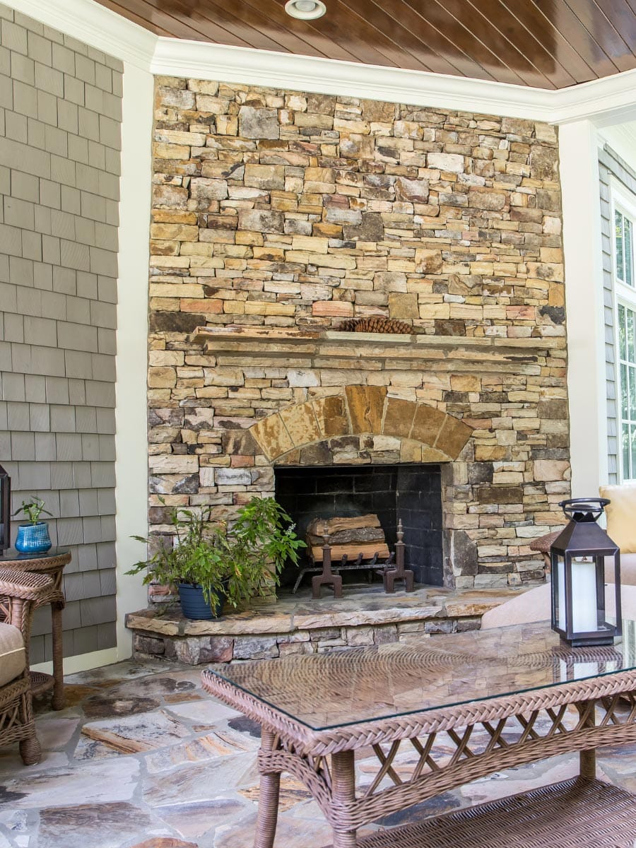Outdoor seating area with wicker furniture in front of a large stone fireplace and chimney. A black lantern and a potted plant stand on one side, with wood paneling and white trim in the background.