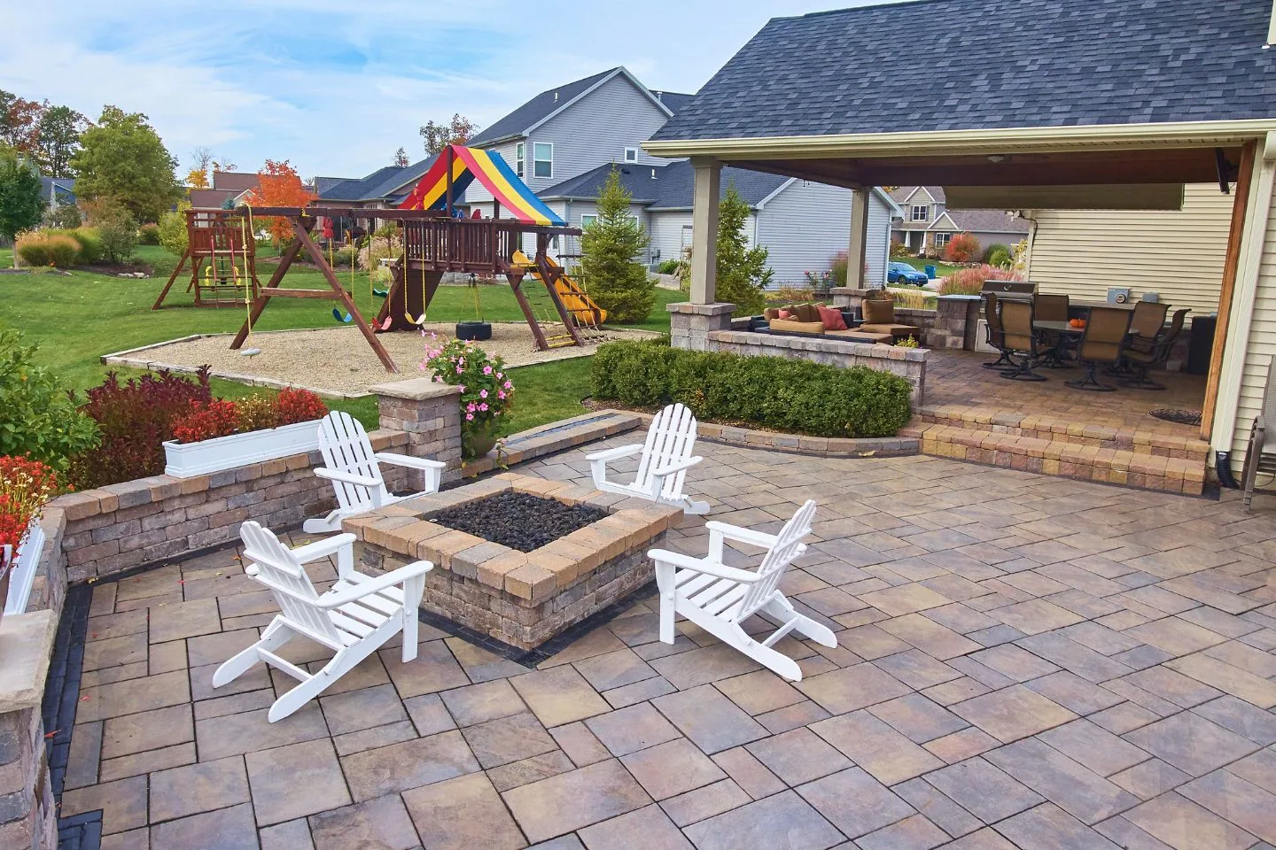 The backyard boasts a tiled patio with fire pits taking center stage, surrounded by four white chairs. A covered outdoor kitchen caters to culinary enthusiasts, while a wooden playground set adds charm to the scene.