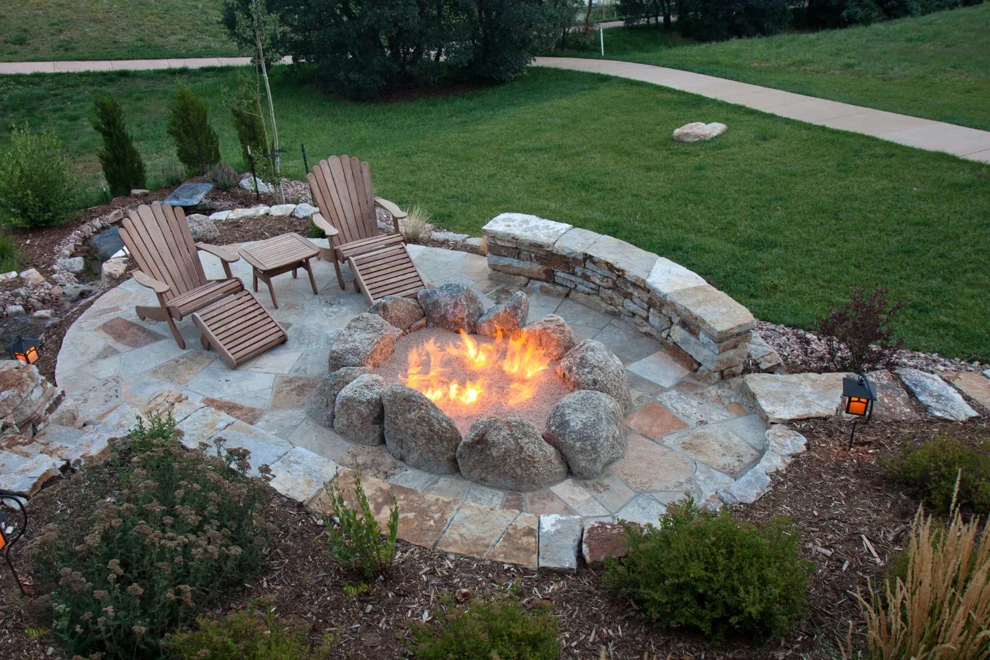 A stone fire pit with a lively flame takes center stage, surrounded by rocks and two wooden chairs on a cozy patio, set amidst a beautifully landscaped garden area.
