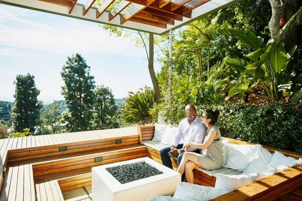 A couple relaxes on a wooden outdoor bench, embraced by lush greenery and enhanced by backyard features like a fire pit in the center, offering warmth and a scenic view in the background.
