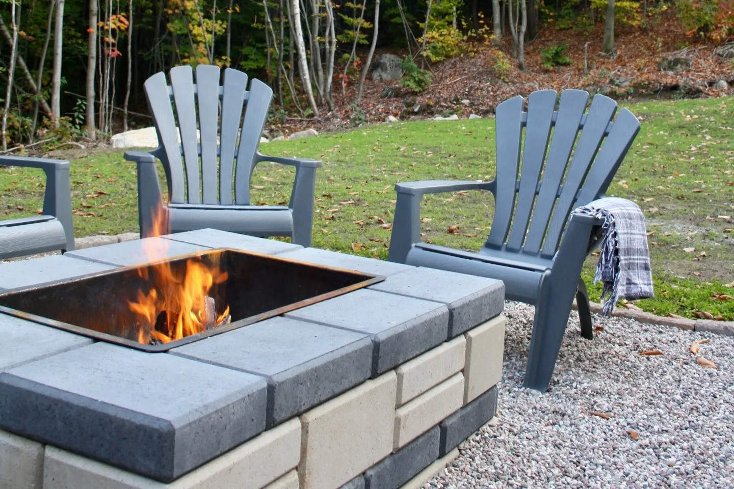 Gray Adirondack chairs encircle a square stone fire pit, where a lively fire dances, set on a gravel patio surrounded by lush greenery and trees. Perfect evenings are crafted here beside one of the finest fire pits.