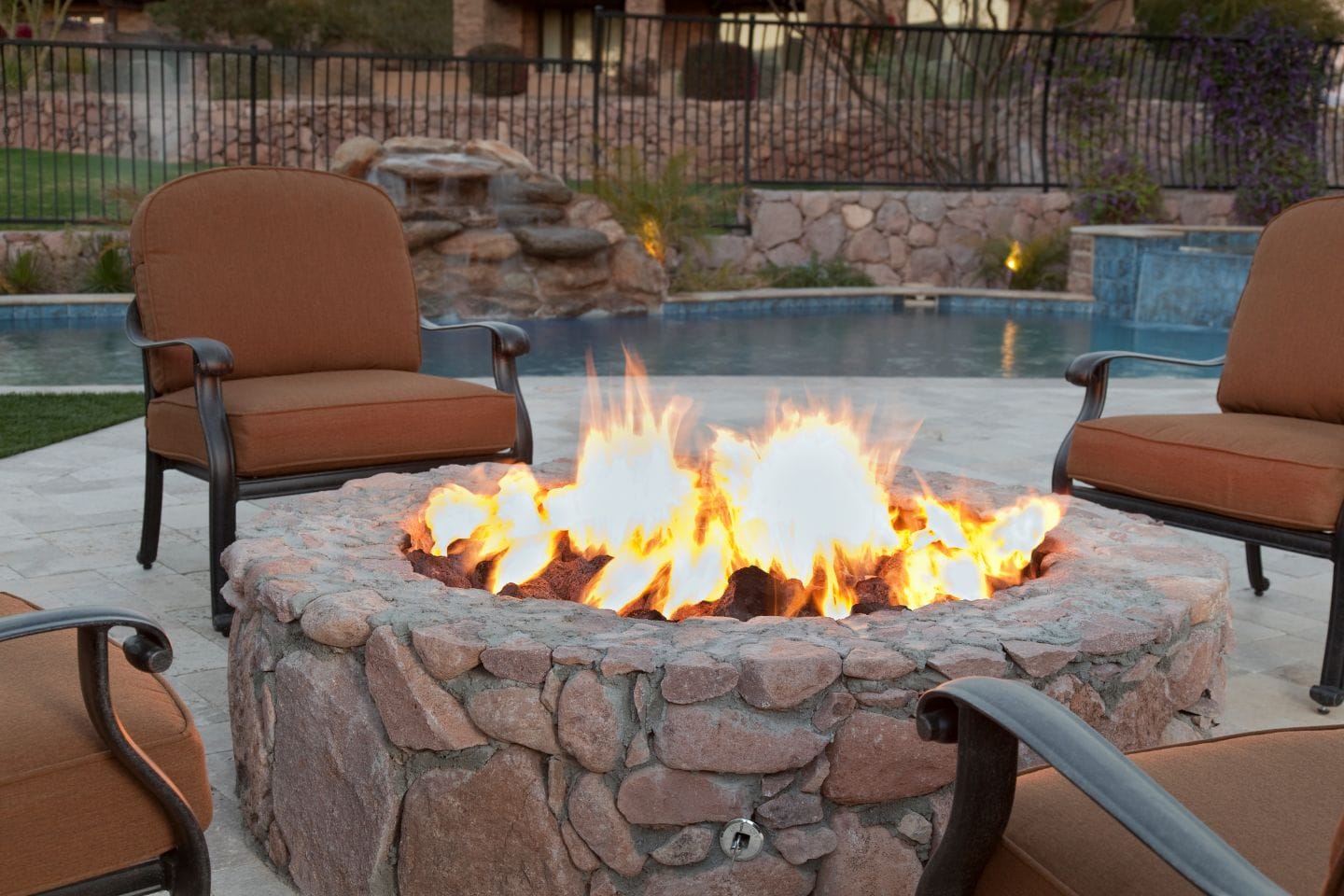 Outdoor stone fire pit with flames surrounded by brown cushioned chairs, creating a cozy garden fire feature by the pool in a backyard setting.