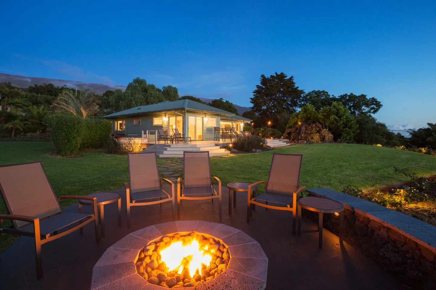 Evening view of a cozy backyard featuring fire pits surrounded by chairs. A house with lit windows stands in the background, and the sky is clear.