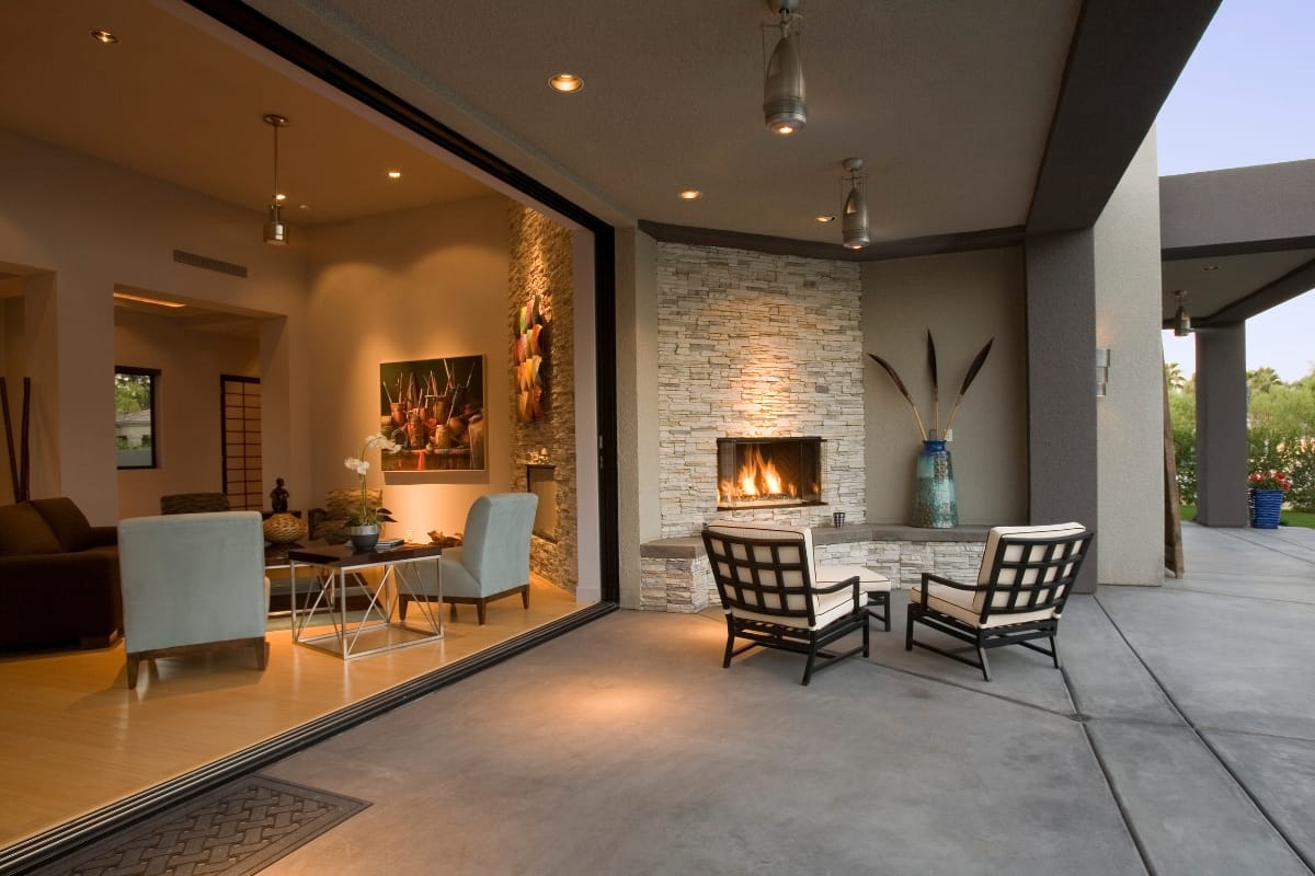 Modern indoor-outdoor living area with an open sliding door, a fireplace, and a table on the patio near a stylish outdoor chimney, with a seating arrangement indoors beside contemporary art.