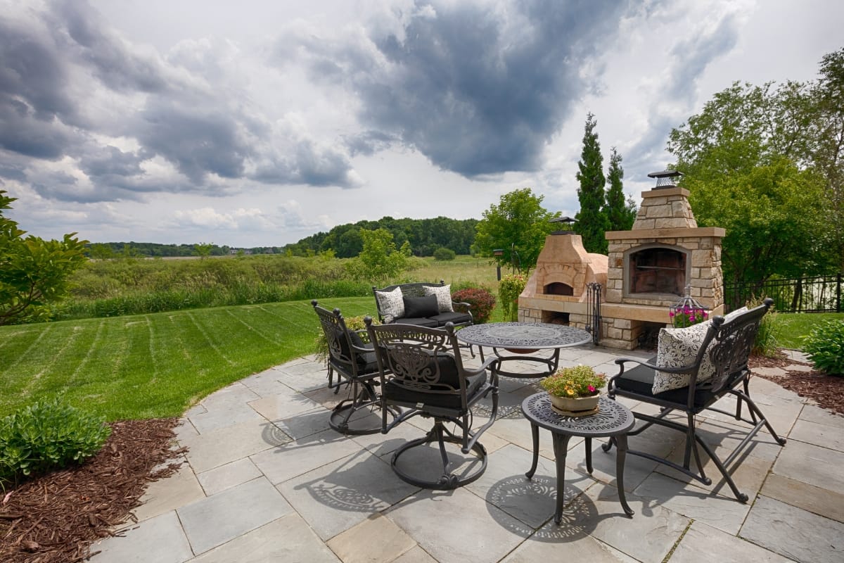 A patio with wrought-iron furniture, including a table and chairs, and a stone outdoor fireplace. The patio overlooks a grassy lawn and a scenic, cloudy landscape, enhanced by the charm of outdoor chimneys.