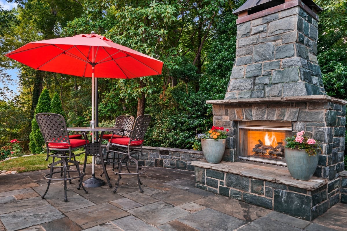 Outdoor patio with a stone fireplace and chimneys, red umbrellas, and a table with four chairs. Surrounded by greenery and potted flowers.