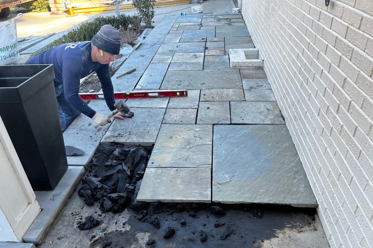 A person is installing rectangular stone tiles on the ground next to a beige brick wall, using a red leveling tool to ensure even placement. Tools and materials are scattered around the work area, typical of patio construction.
