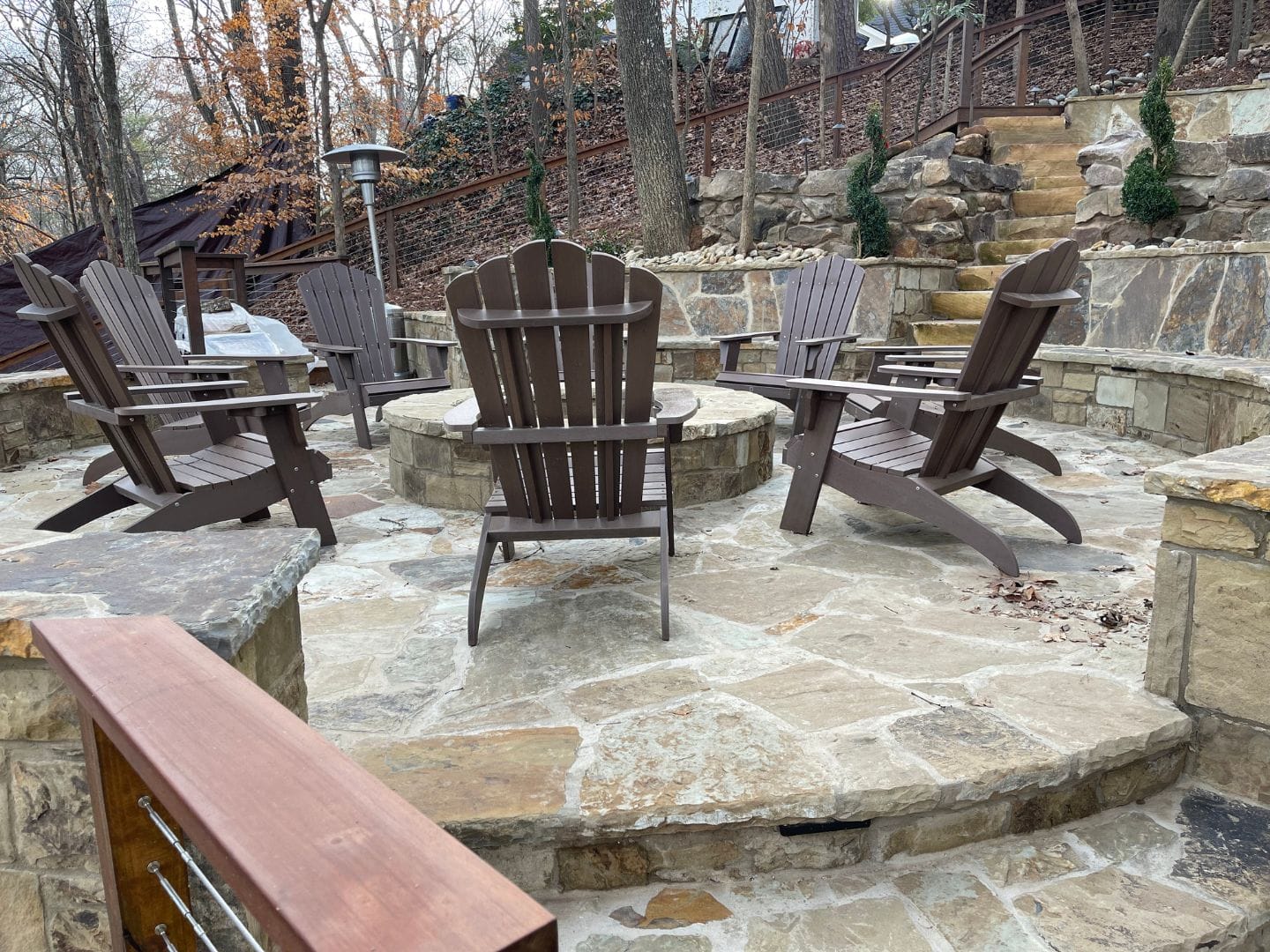 Outdoor living areas: A stone patio with Adirondack chairs arranged in a circle around a central fire pit. The area is surrounded by exquisite garden spaces and tall trees in the background, creating an inviting outdoor oasis.
