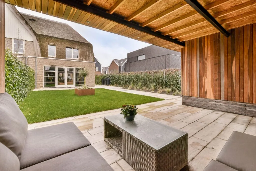 A custom patio design features a covered space with a gray sofa and woven table, overlooking a grass lawn and two-story houses with large windows.