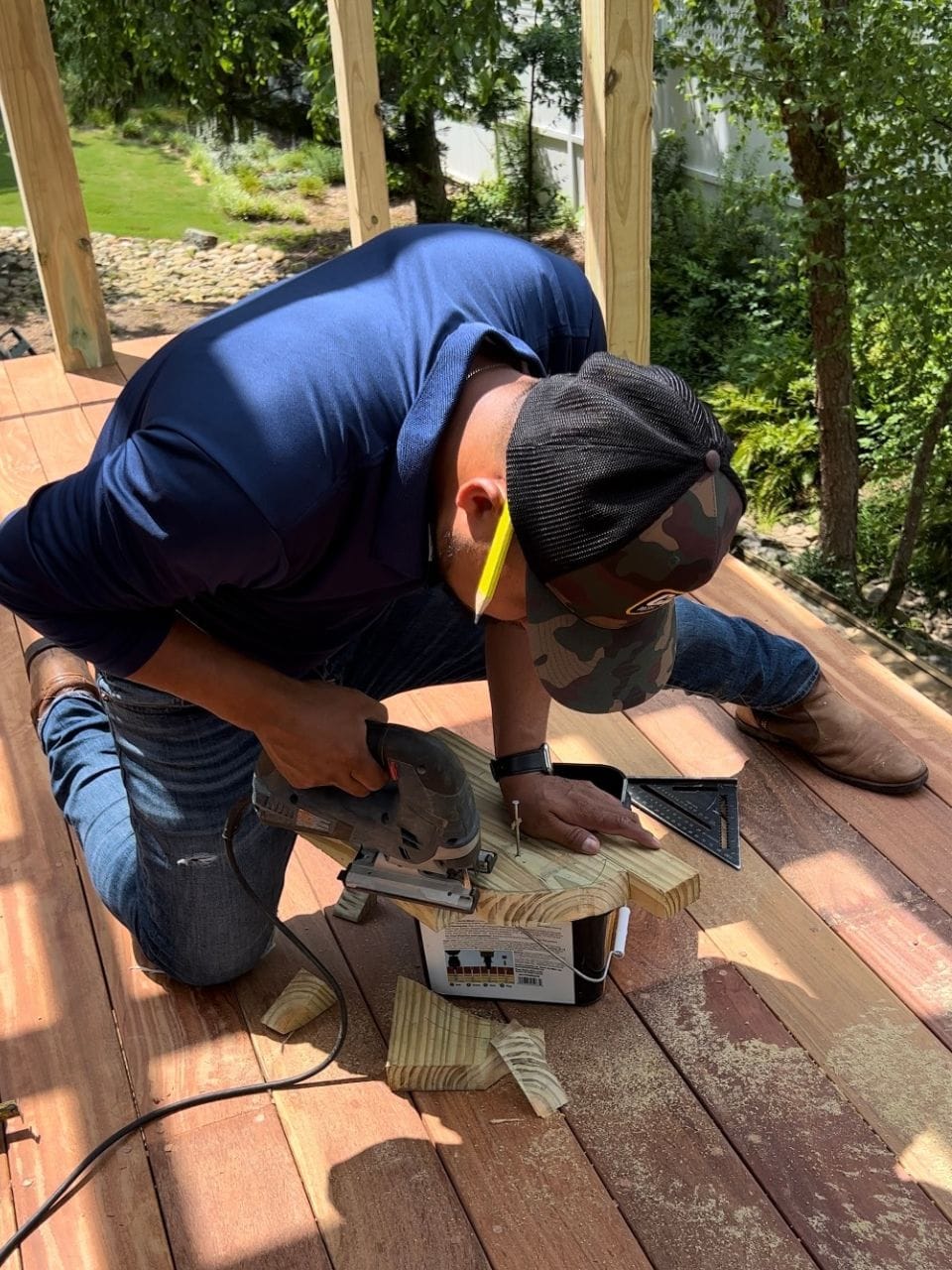 A person wearing a cap and safety glasses uses a circular saw to cut a piece of wood on a deck, likely preparing for outdoor patio improvements. Sawdust is visible around the cutting area.