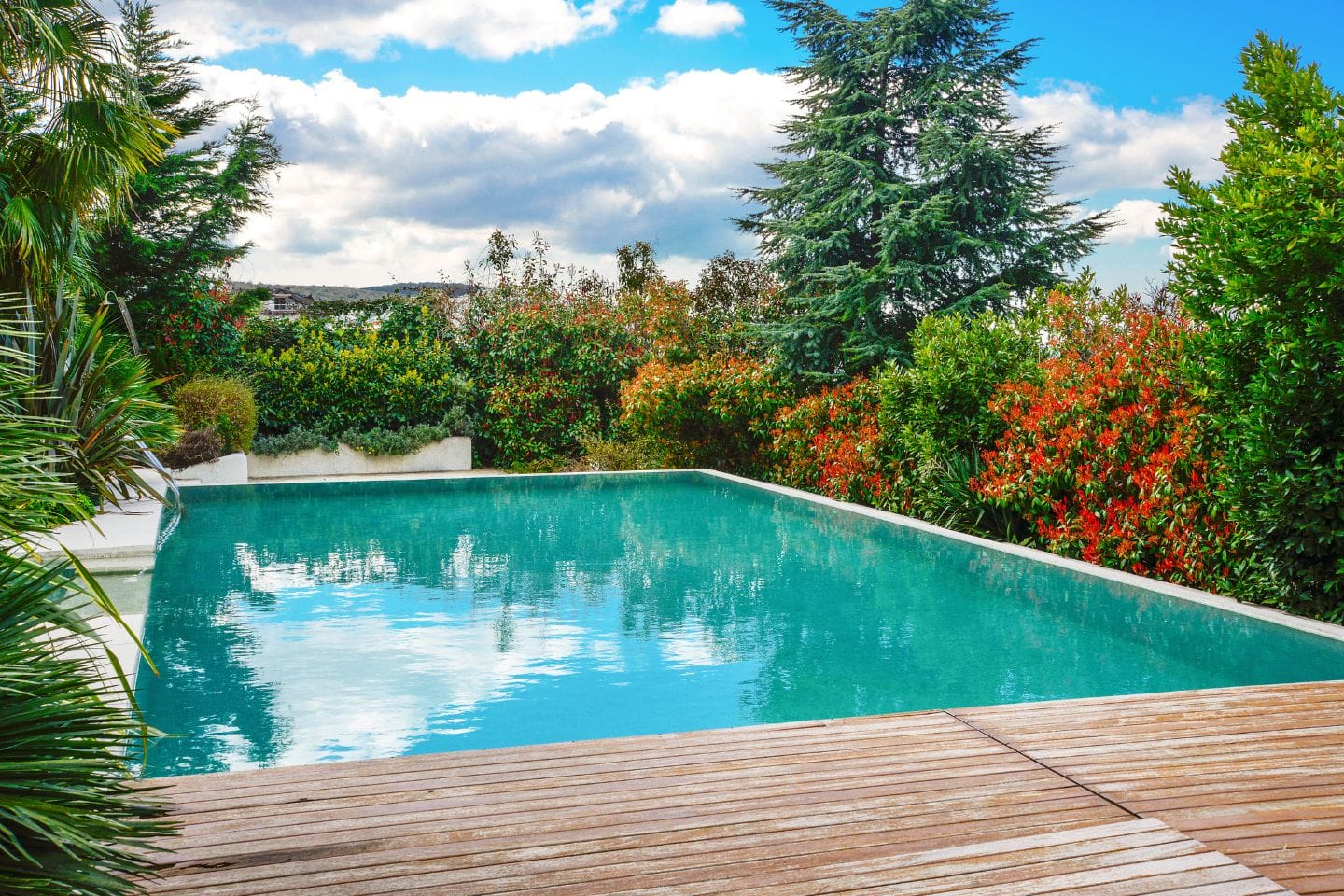 A rectangular outdoor swimming pool, fresh from recent renovations, is surrounded by trees and bushes. A wooden deck stretches invitingly in the foreground under a partly cloudy blue sky.