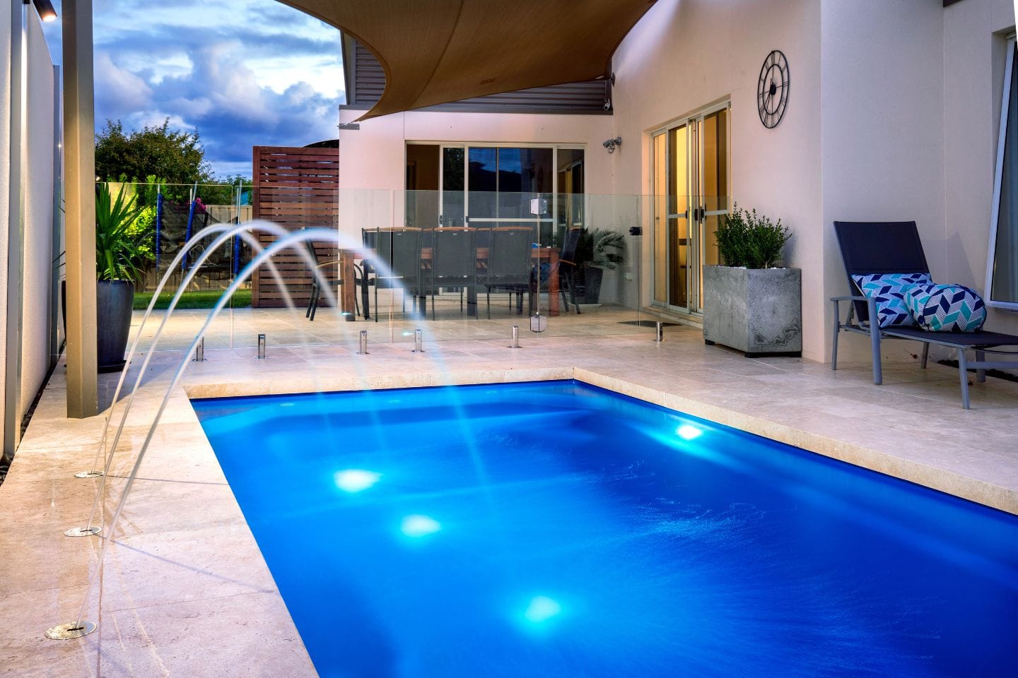 Modern backyard with a small, illuminated pool and a fountain, showcasing expert pool renovations. Patio area with glass fencing, dining table, and lounge chairs under a shade sail offers the perfect blend of style and relaxation.