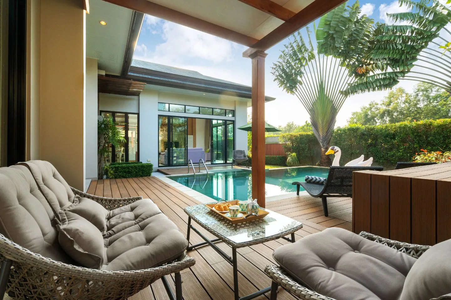 An outdoor patio with cushioned wicker chairs and a small table overlooks a pool, recently refreshed through pool renovations. A swan float gently drifts in the water, surrounded by lush plants and towering trees that enhance the serene atmosphere.