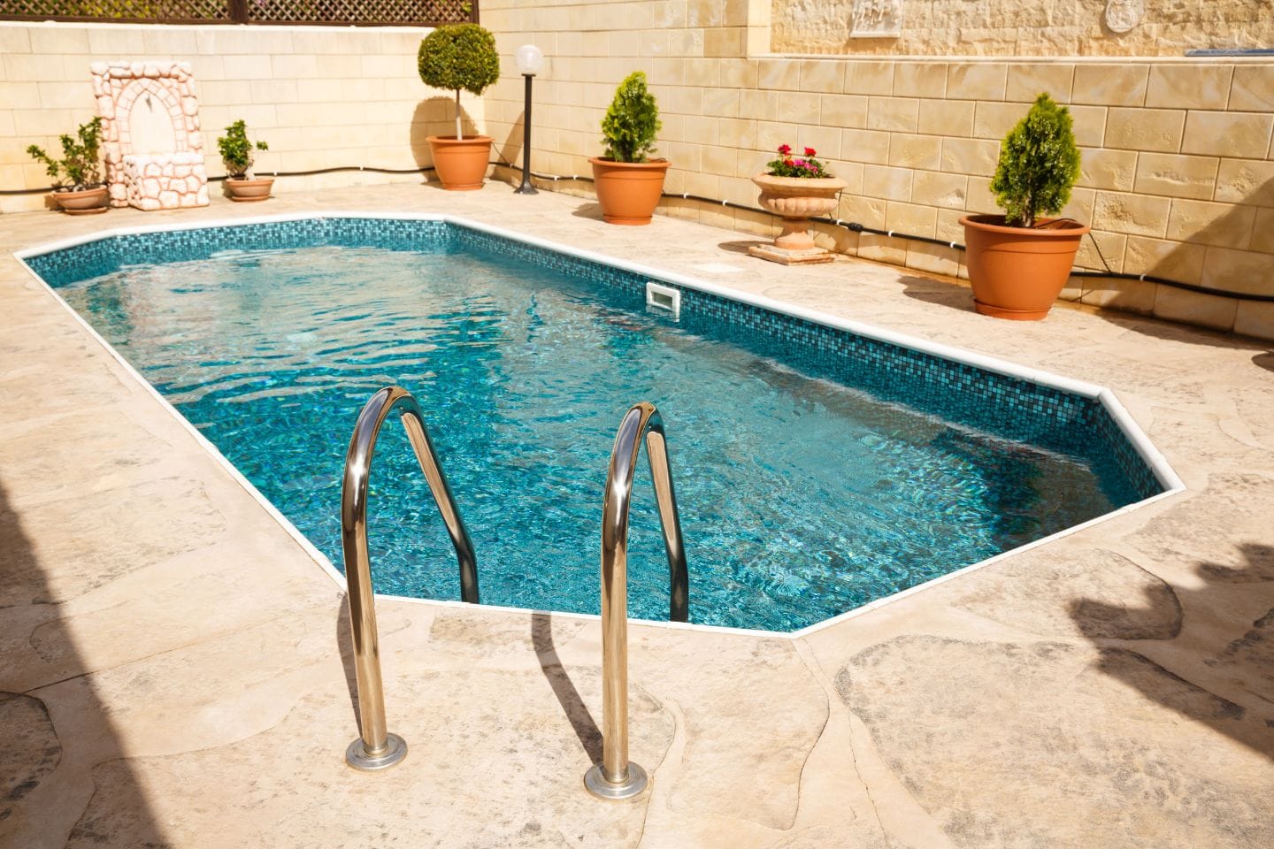 A small, blue outdoor swimming pool with metal steps is surrounded by a stone patio, showcasing recent pool renovations. Several potted plants are thoughtfully arranged around the pool for a touch of nature.