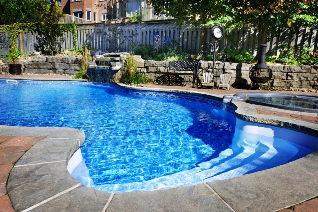 Curved swimming pool with clear blue water, surrounded by stone paving. A fence, plants, and seating area with a small waterfall feature are in the background—a perfect spot for those considering swimming pool upgrades.