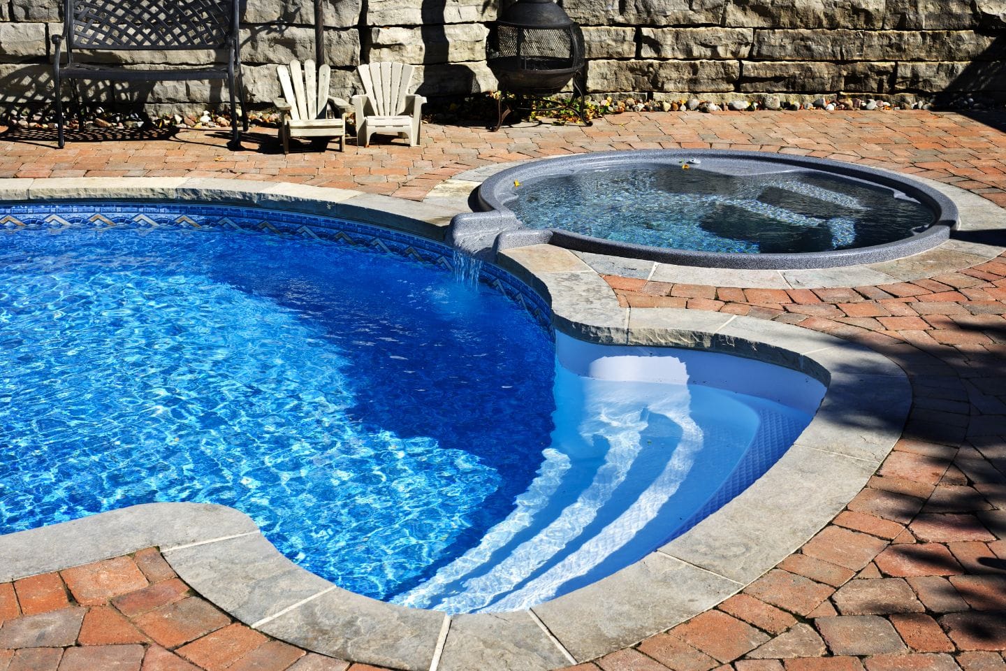 A curvy swimming pool and a connected round hot tub with clear blue water, the results of recent pool renovations, are surrounded by a brick patio. Two white chairs and a metal fireplace sit in the background near a stone wall.