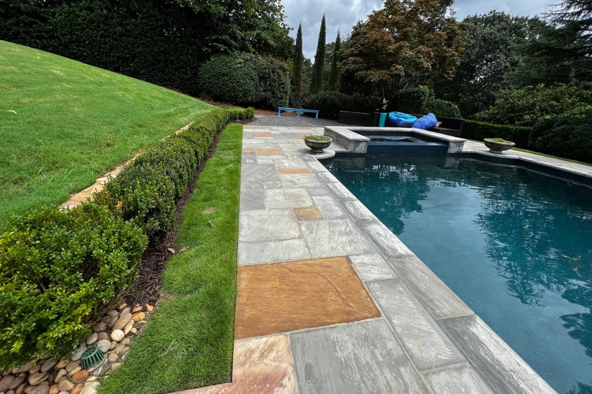 A backyard pool area with stone paving, green lawn, trimmed hedges, and a few poolside lounge chairs and a table in the background. The recently completed swimming pool updates include two planters at one end.