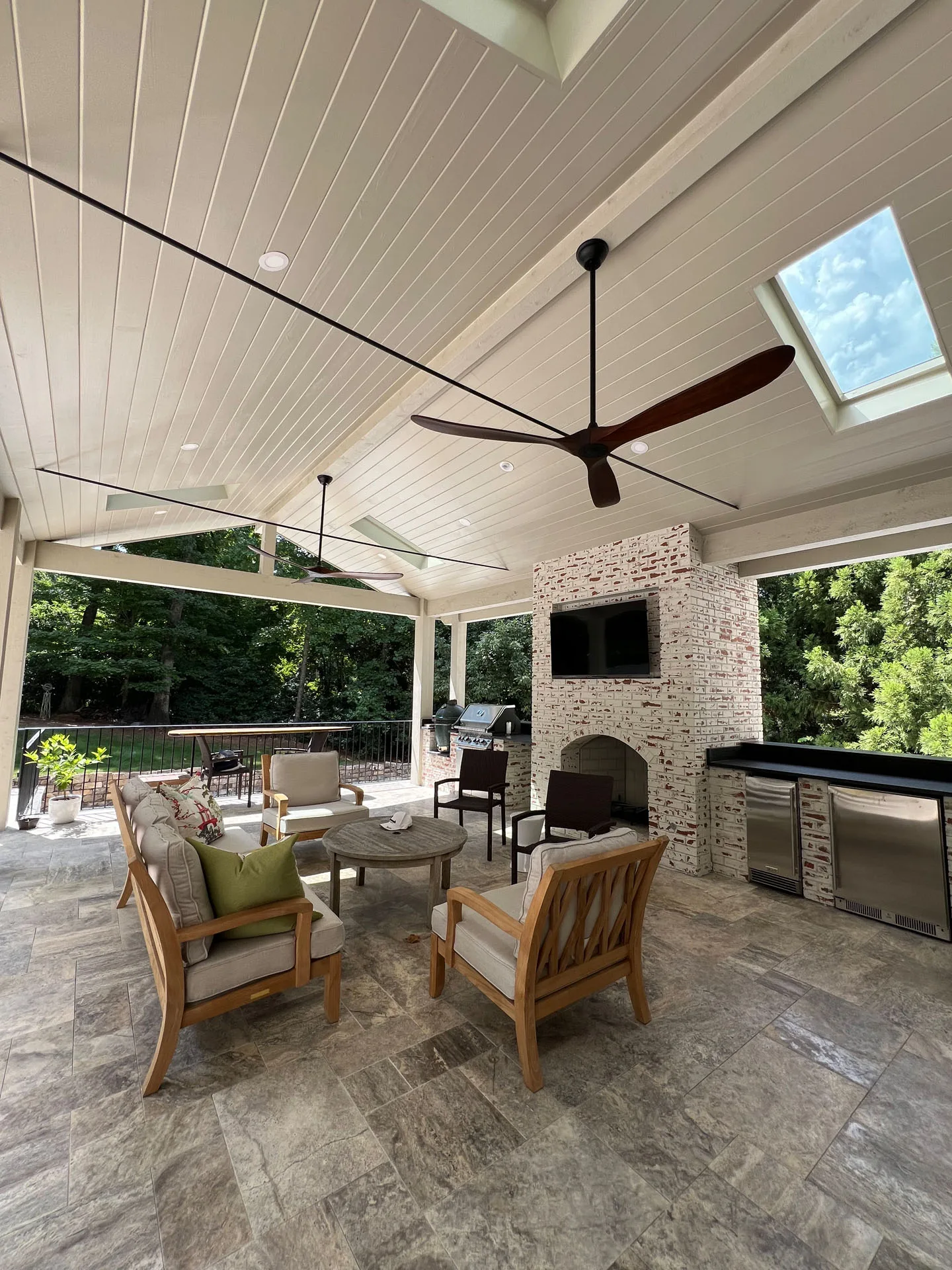 Covered outdoor patio with seating, a ceiling fan, skylights, television, and outdoor kitchen area. Surrounded by trees, it has a modern stone floor and a partially brick wall.