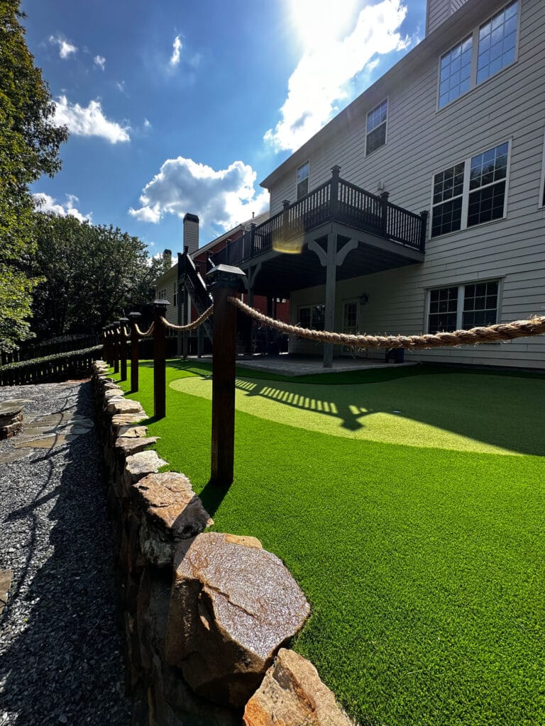 A backyard with artificial turf, bordered by a stone wall and rope fence. A two-story house with a wooden deck is in the background under a blue sky with clouds.