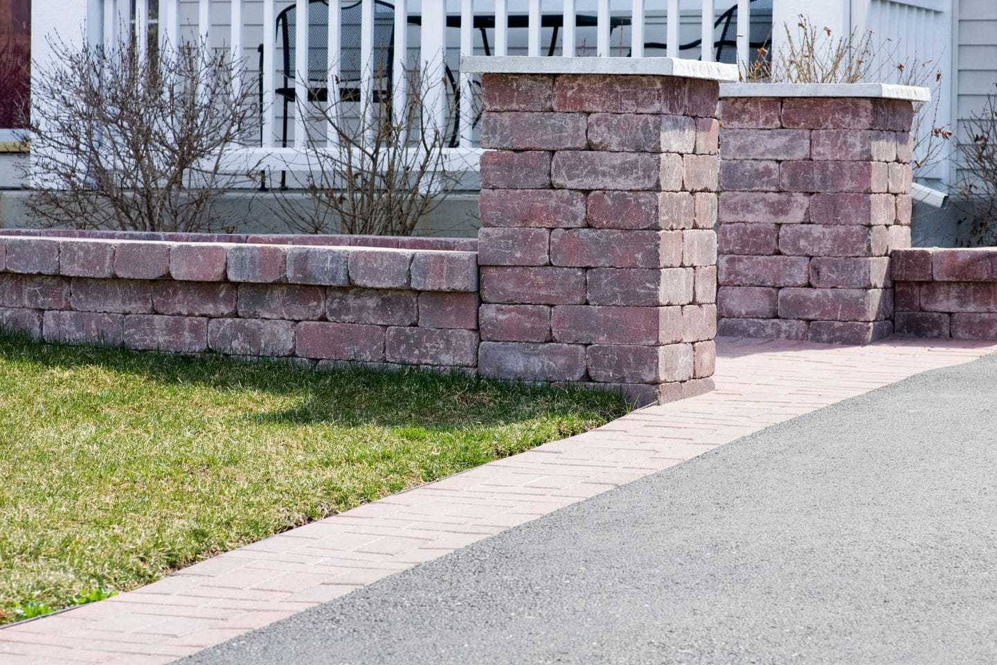 Brick pillars and a low wall, expertly crafted by retaining wall contractors, define a yard next to a concrete driveway. Sparse bushes and part of a white porch are visible in the background.