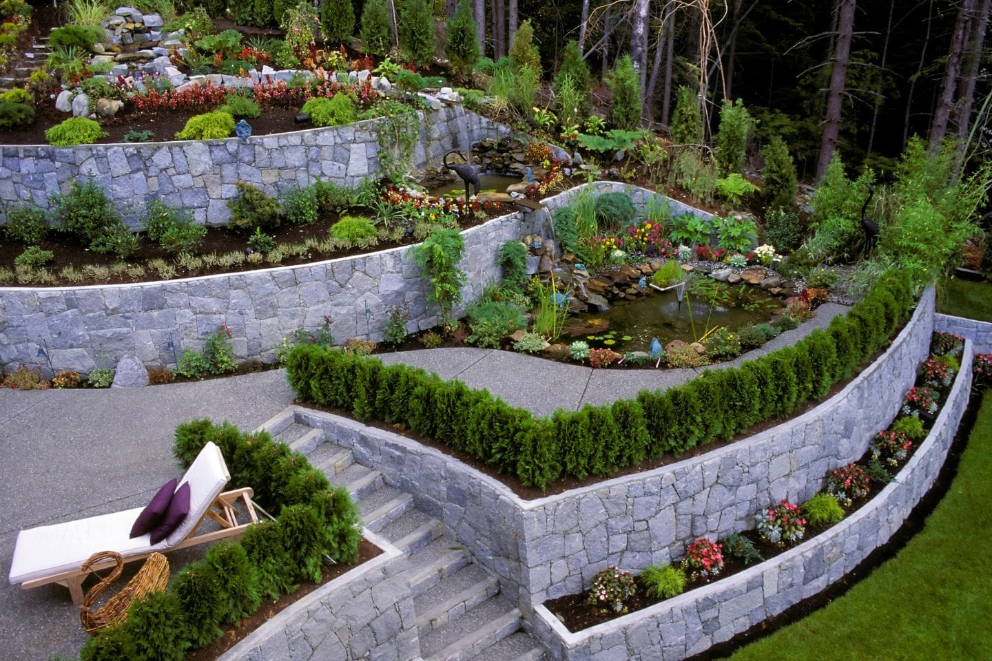 Terraced garden with expertly crafted stone walls by retaining wall contractors, lush shrubs, colorful flowers, and a tranquil pond. Gray steps lead to a cozy lounge chair with cushions in the foreground.