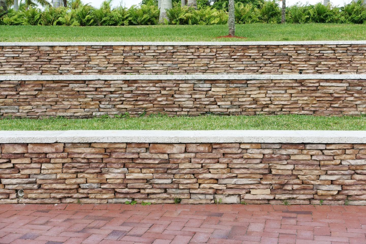 Experienced retaining wall contractors have crafted terraced stone retaining walls with patches of grass in between, surrounded by lush greenery.