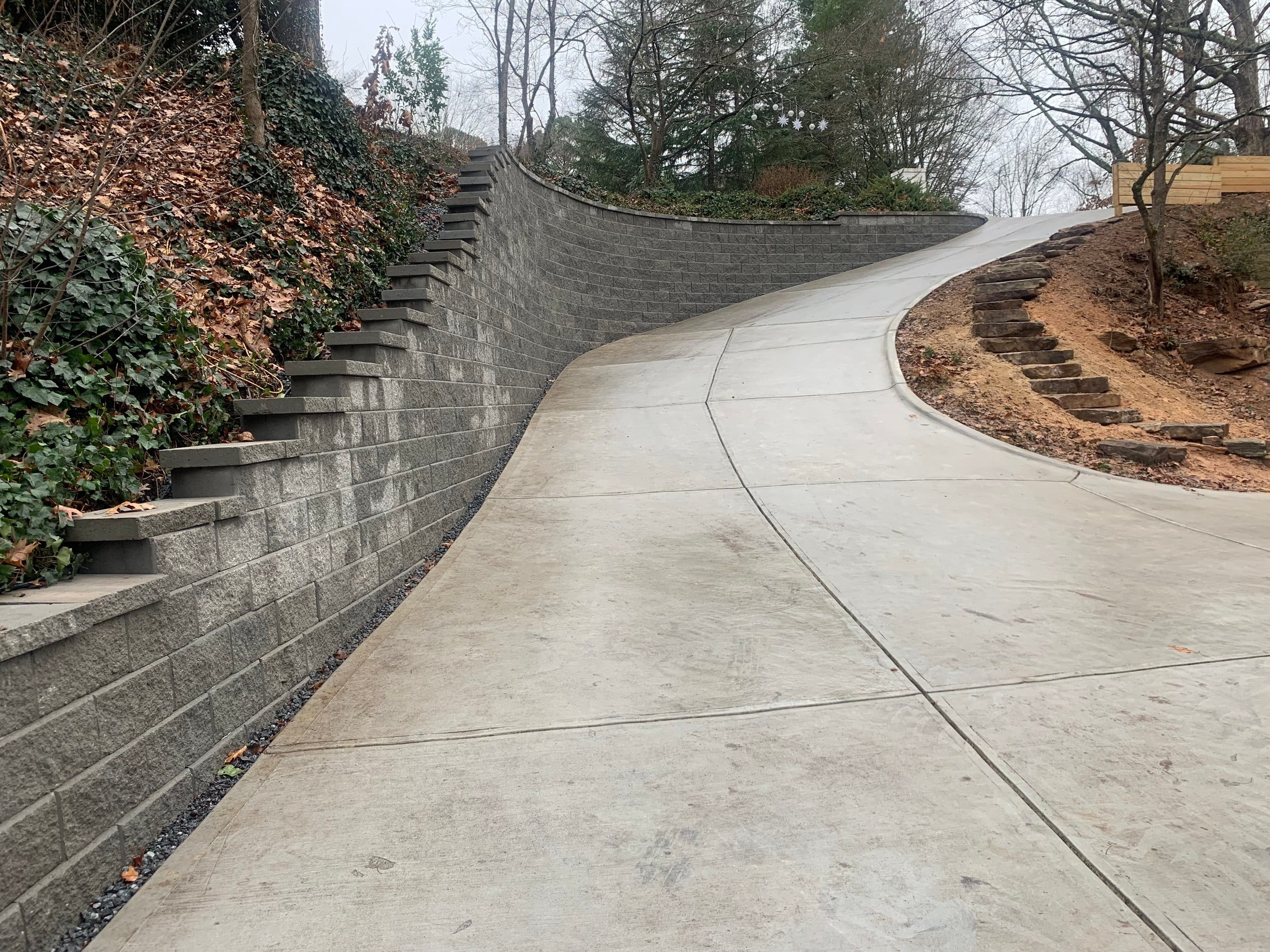 A steep, smoothly paved concrete driveway lined with expertly-crafted retaining walls on both sides, surrounded by trees and foliage, showcasing impeccable hardscaping.