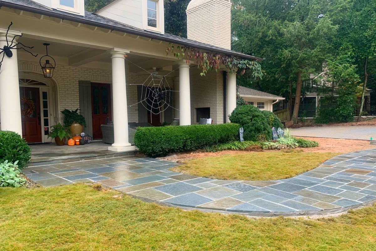 A house with a decorated porch featuring columns, a spider web, pumpkins, and a tile walkway leading to the front door. Greenery and grass surround the area. A small building is visible in the background, along with well-maintained sidewalks weaving through the lush lawn.