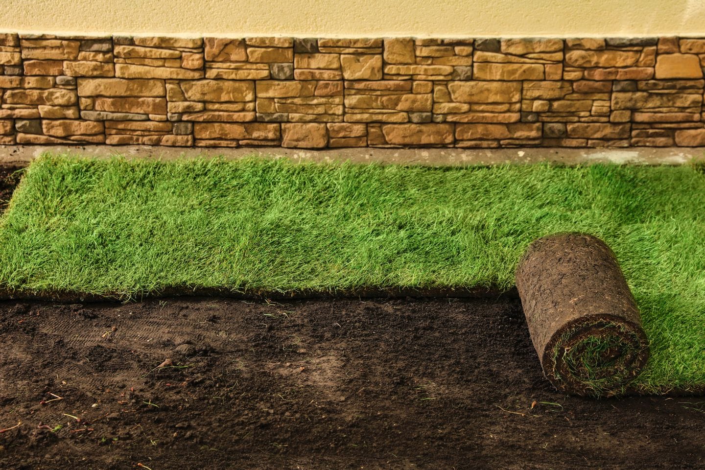 A roll of sod partially unrolled on dark soil, with a section already laid, complements the expertly crafted stone wall in the background, highlighting professional hardscaping services.