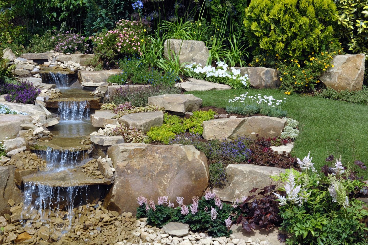 A landscaped garden featuring a small cascading waterfall, bordered by rocks and lush greenery, with colorful flowers and shrubs scattered around—an oasis that offers more than just beauty, akin to premium SEO services for your outdoor space.