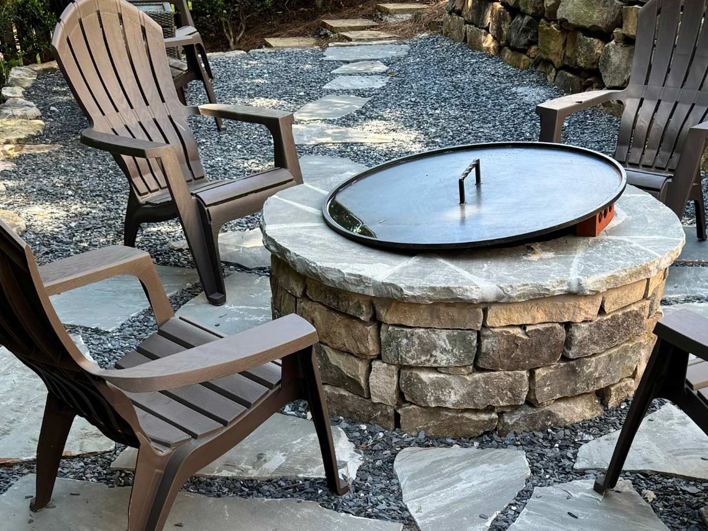 A circular fire pit with a metal cover is surrounded by four brown Adirondack chairs, creating a cozy gathering spot on the stone patio.