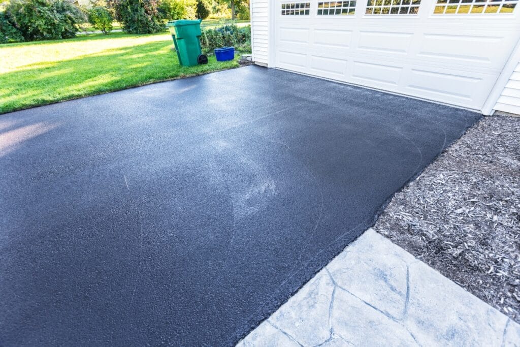 Freshly paved black asphalt driveway in front of a white garage, with green lawn, recycling bin, and shaded trees in the background—perfect for those seeking modern driveway paving ideas.