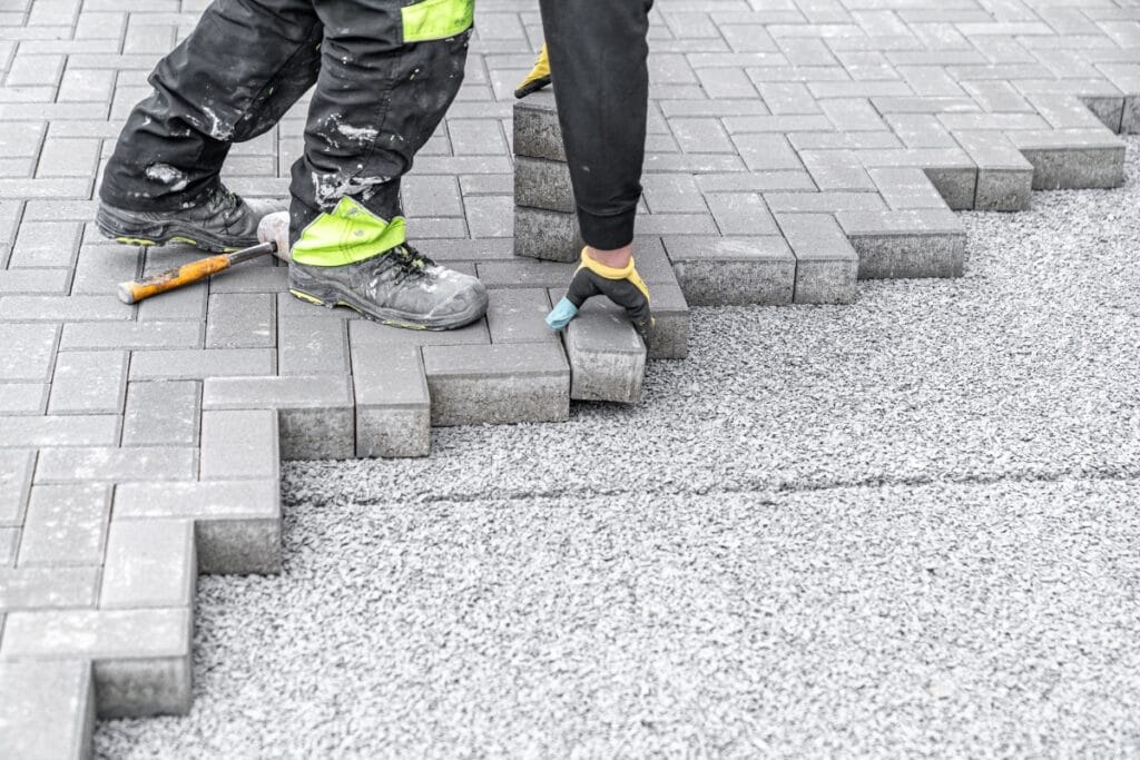 A worker places rectangular concrete pavers to create a patterned pavement, showcasing practical driveway paving ideas, with tools and gravel visible nearby.