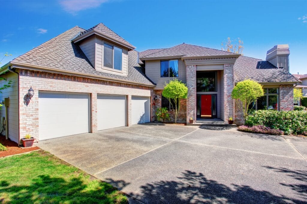Two-story suburban house with a brick and siding exterior, three-car garage, large driveway showcasing modern driveway paving ideas, and landscaped front yard under a clear blue sky.