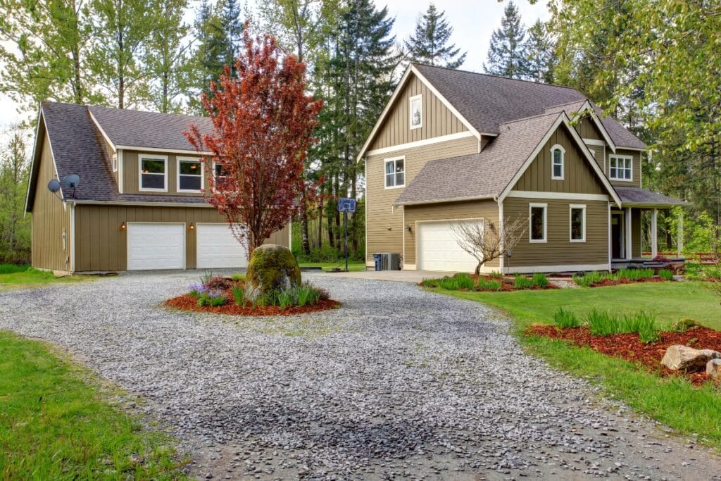 Two beige houses with white trim and brown roofs, separated by a gravel driveway, surrounded by trees and landscaped greenery—an inviting scene perfect for exploring new driveway paving ideas.