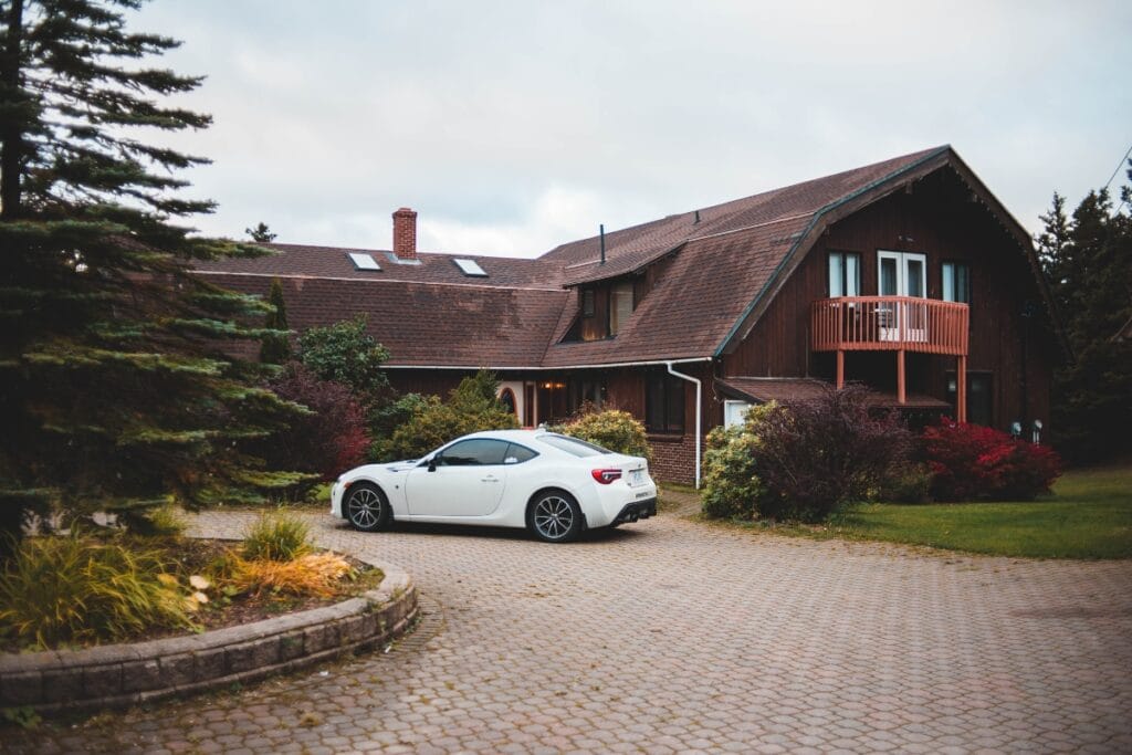 A white sports car is parked on a paved driveway in front of a two-story house with a sloped roof and small balcony, showcasing stylish driveway paving ideas.