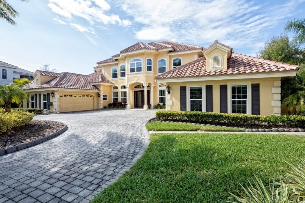 Two-story yellow stucco house with red tile roof, three-car garage, large windows, manicured lawn, and a paved driveway—perfect for showcasing your favorite driveway paving ideas—under a partly cloudy sky.