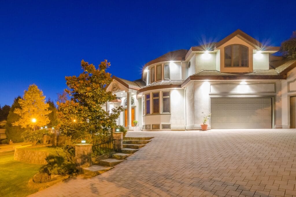 Large, modern two-story house with a curved driveway, three-car garage, and bright exterior lighting, photographed at dusk—perfect for those seeking elegant driveway paving ideas.
