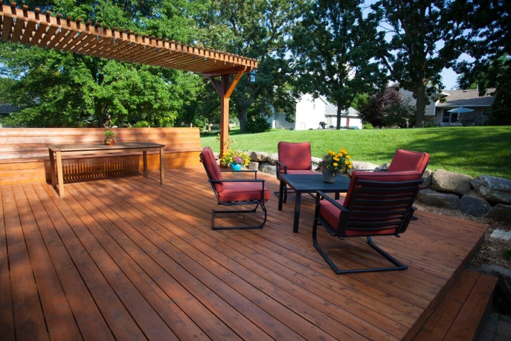 A wooden patio with four red cushioned chairs, two black tables, a wooden pergola, and bench seating overlooks lush grass and trees—a perfect spot to relax while keeping deck maintenance in mind.