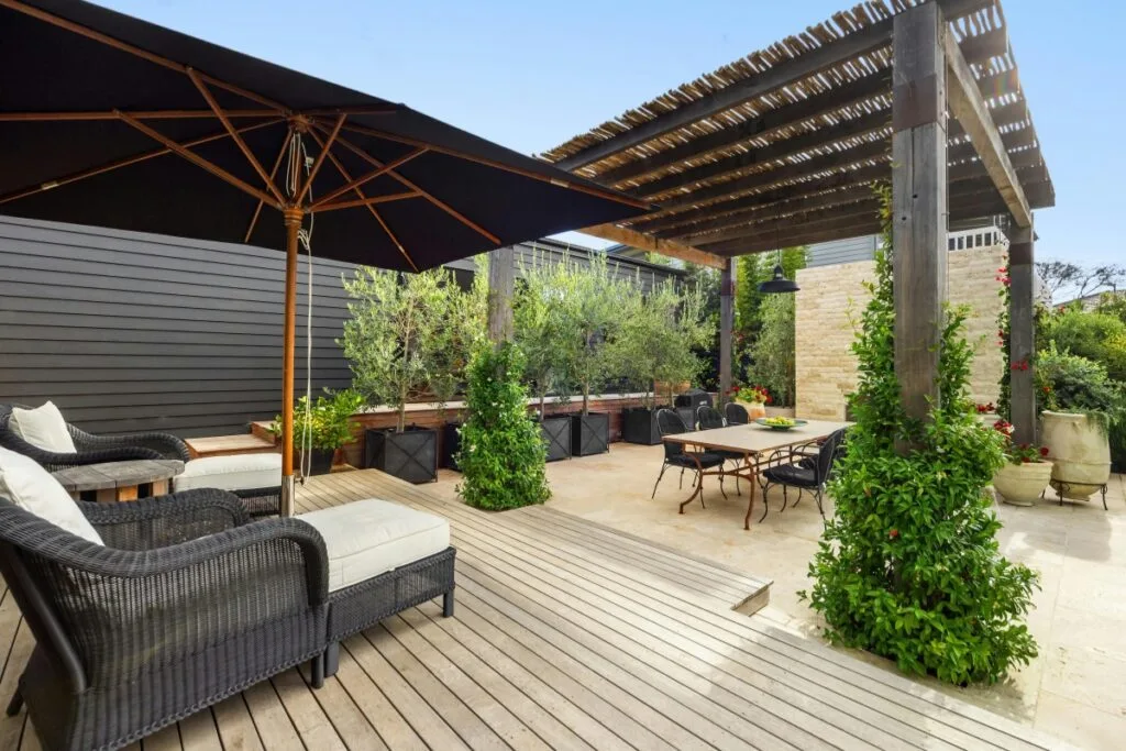 Modern outdoor patio with wicker seating, a large black umbrella, wooden deck featuring easy deck maintenance, dining table, pergola with slats, and greenery including potted plants and climbing vines.