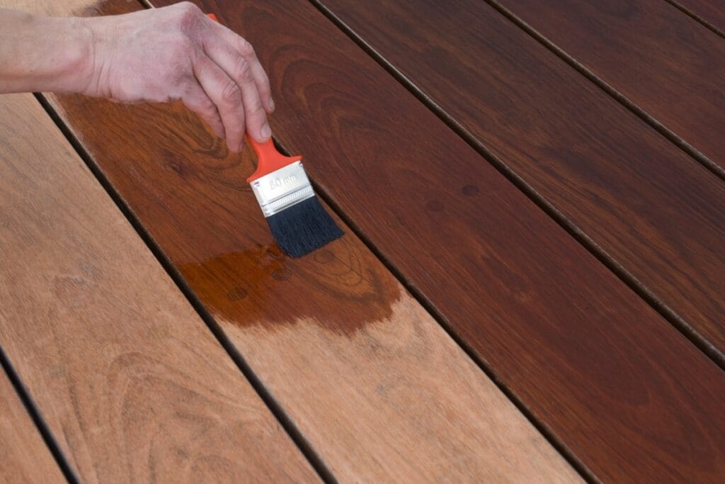 A hand uses a paintbrush to apply wood stain to a wooden deck, changing the surface from light brown to a darker finish as part of regular deck maintenance.
