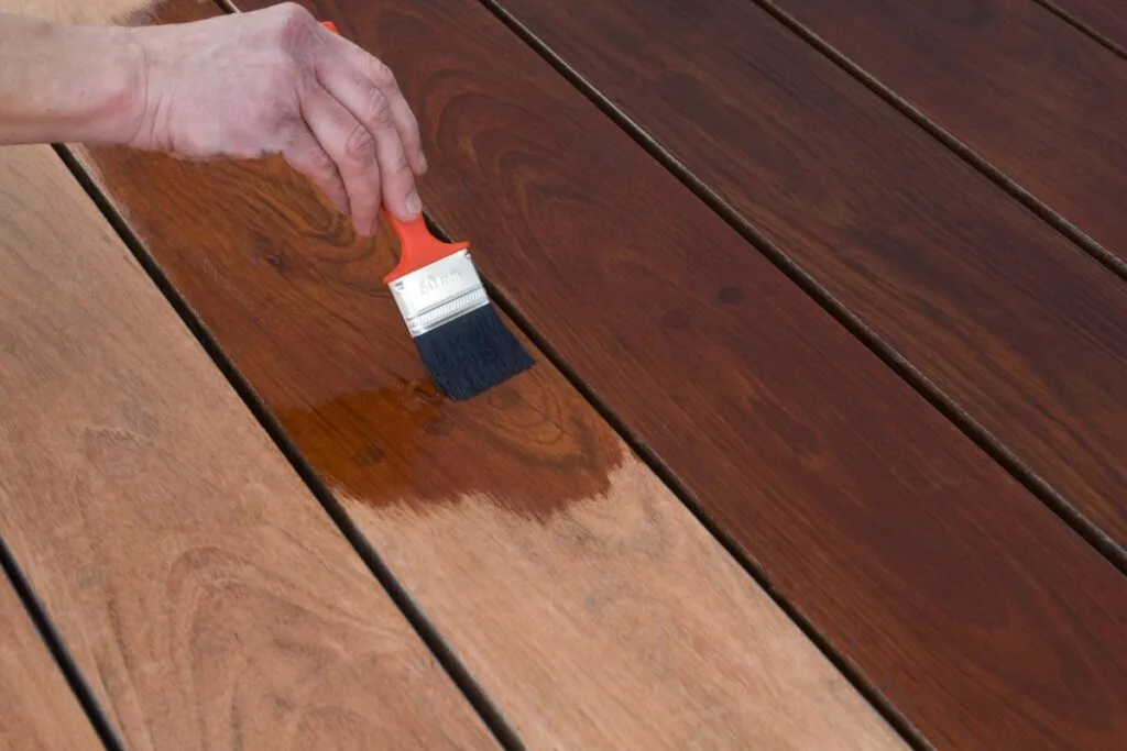 A hand uses a paintbrush to apply wood stain to a wooden deck, changing the surface from light brown to a darker finish as part of regular deck maintenance.