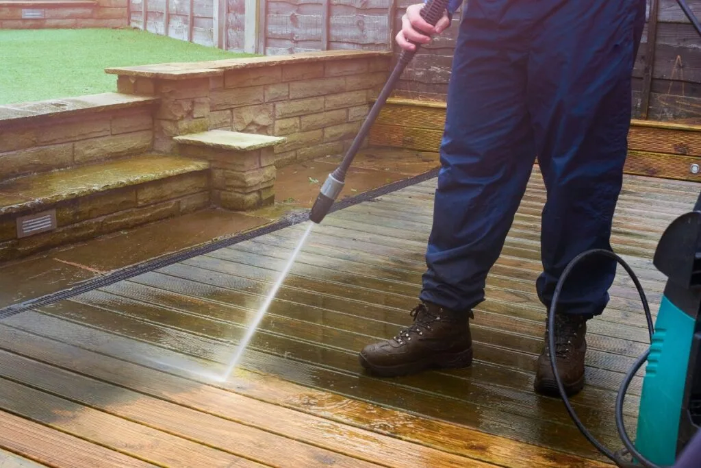 A person focuses on deck maintenance outdoors, using a pressure washer to clean a wooden deck while dressed in blue pants and brown boots.