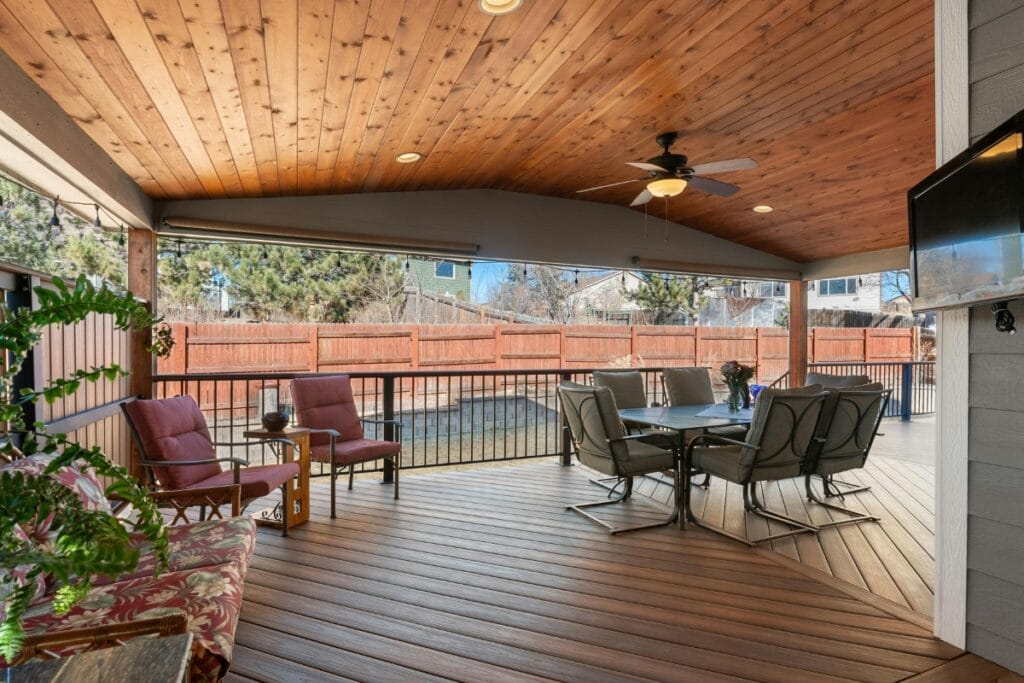 Covered patio with wood ceiling and floor, outdoor dining set, armchairs, potted plants, ceiling fan, mounted TV, and a backyard view—ideal for relaxing or entertaining. Easy deck maintenance keeps this inviting space looking its best year-round.