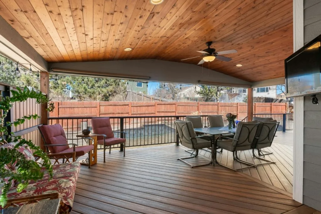Covered patio with wood ceiling and floor, outdoor dining set, armchairs, potted plants, ceiling fan, mounted TV, and a backyard view—ideal for relaxing or entertaining. Easy deck maintenance keeps this inviting space looking its best year-round.