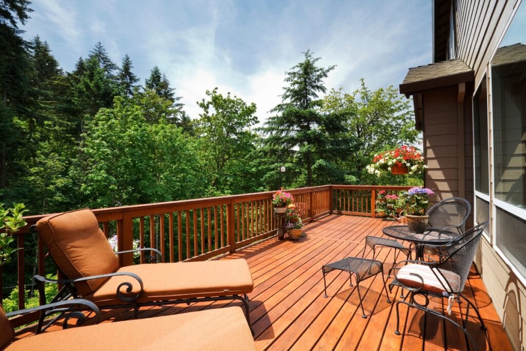 A wooden deck with patio furniture, potted flowers, and lounge chairs overlooks a lush green forest under a partly cloudy sky—the perfect spot to relax and enjoy easy deck maintenance.