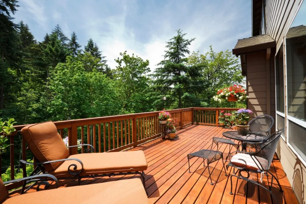 A wooden deck with patio furniture, potted flowers, and lounge chairs overlooks a lush green forest under a partly cloudy sky—the perfect spot to relax and enjoy easy deck maintenance.