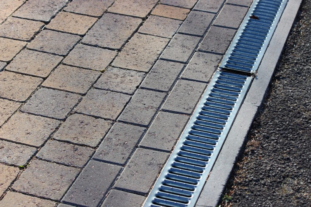 Close-up of a paved walkway made of rectangular bricks next to a metal drainage grate, ensuring proper drainage, with some asphalt visible on the right side.