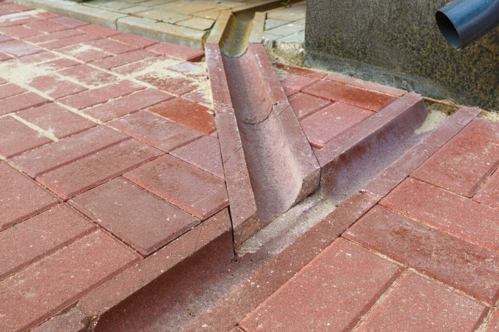 A rainwater drainage channel made of red paving bricks crosses a sidewalk, providing proper drainage by directing water away from a building’s downspout.